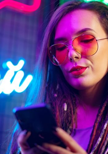Fashion girl using her smartphone while standing near neon signs in nightclub close up
